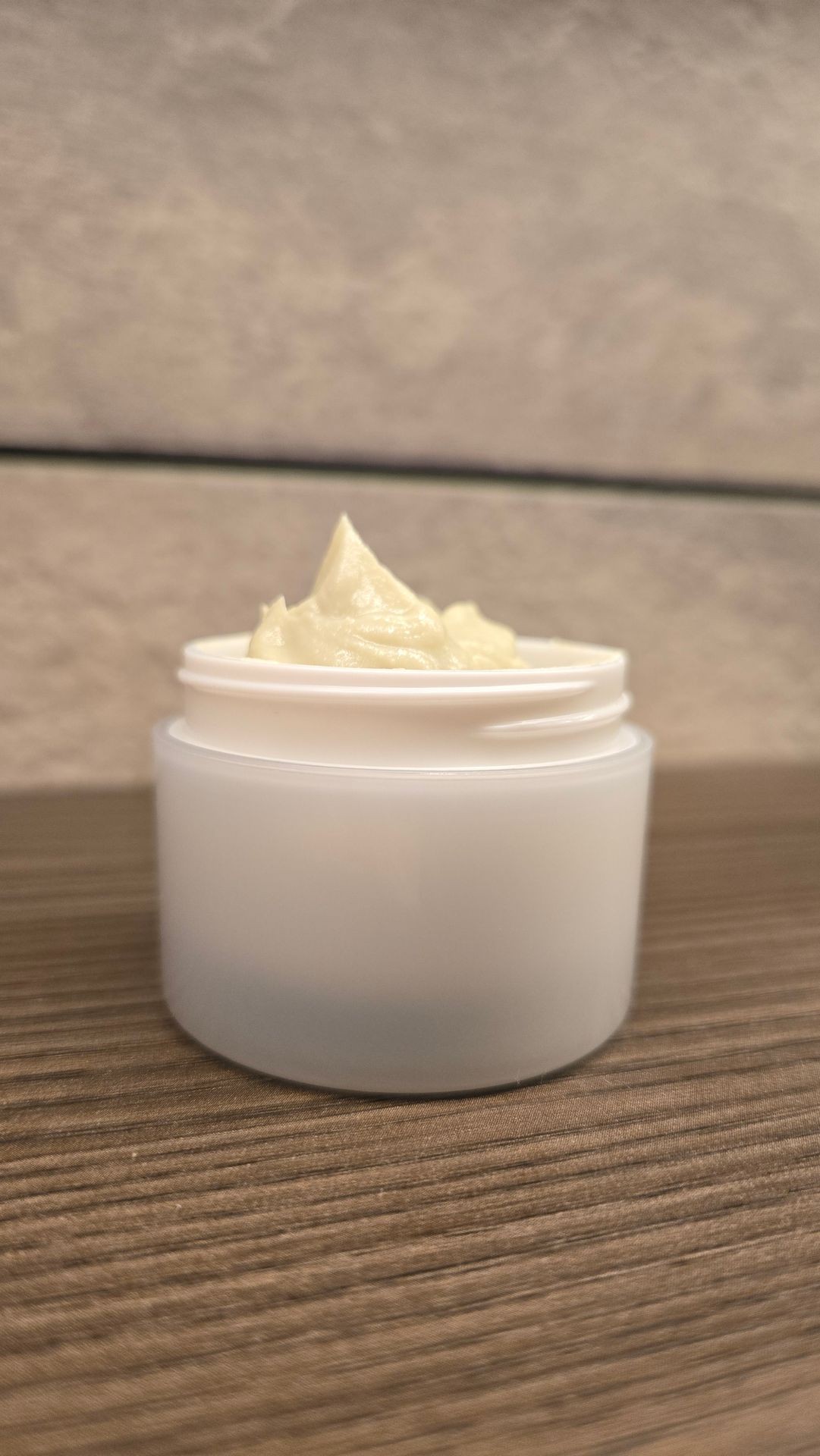 White plastic jar filled with beige cream, sitting on a wooden surface with a blurry stone background.