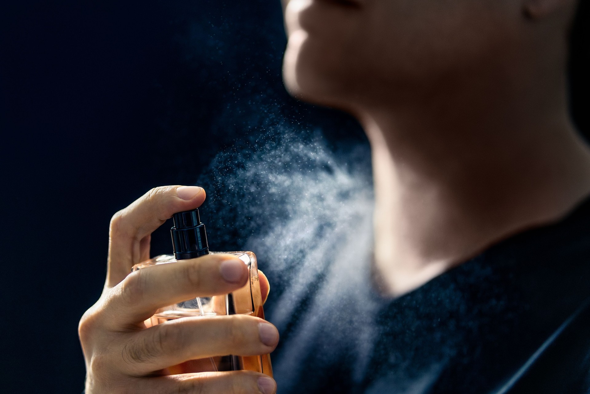 Fragrance spray. Man and perfume. Applying cologne. Scent water. Skin care, beauty product and male cosmetics concept. Holding elegant smell bottle in hand. Dark blue background.