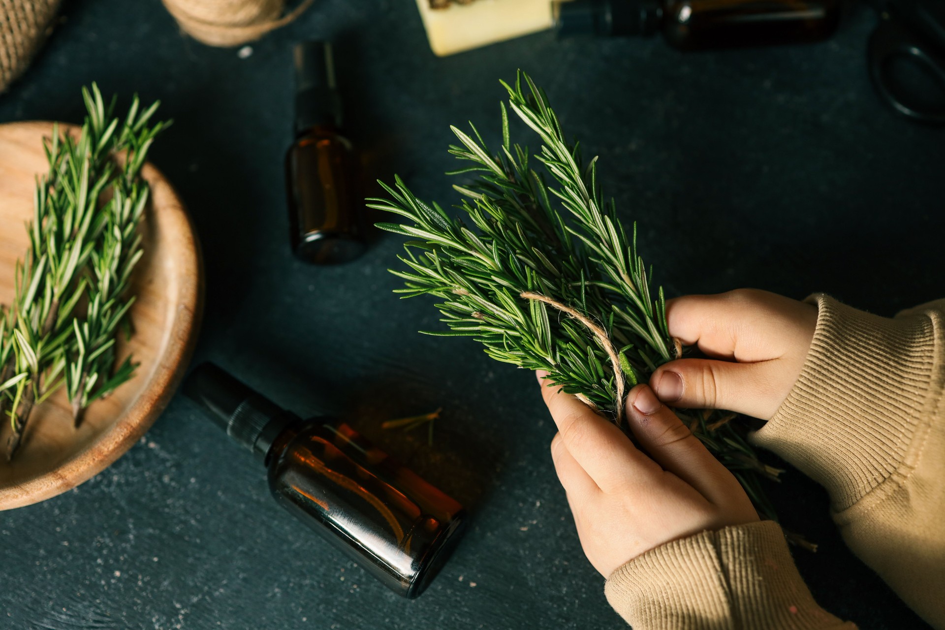 Hands holding a bundle of fresh rosemary tied with twine, surrounded by amber glass bottles, natural soap, burlap fabric, and crafting tools on a dark rustic surface